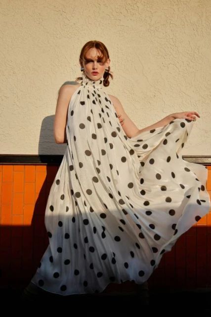 A woman poses outdoors in a long white halter dress with black polka dots, with a neutral background in the sunlight.