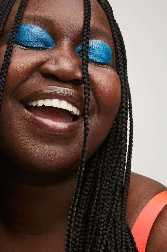 Close-up of a Black woman with braids smiling, blue eyeshadow, and coral top against a light background in a portrait-oriented shot.