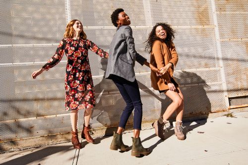 Three women joyfully walk arm in arm down a sunny city street, showcasing casual-chic fashion and footwear. The composition highlights movement and style.
