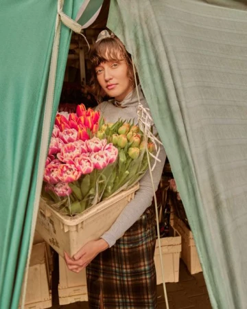 A woman stands holding a crate of colorful tulips, framed by a green tent, capturing a spring flower market scene with natural light.
