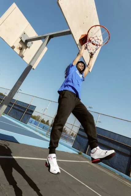 Athlete with dreadlocks performs a slam dunk on an outdoor court in athletic wear and sneakers under clear blue skies during daytime.