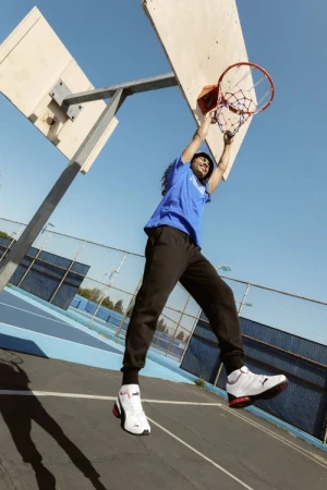 Athlete with dreadlocks performs a slam dunk on an outdoor court in athletic wear and sneakers under clear blue skies during daytime.