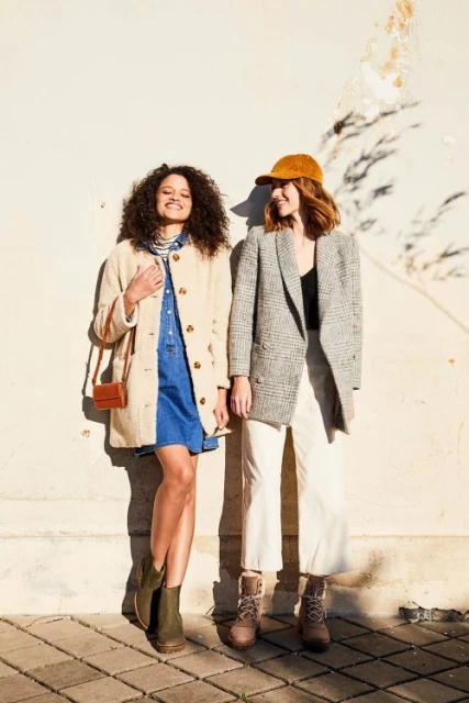 Two young women stand side-by-side against a textured cream wall in natural light, wearing stylish coats, dresses, boots, and accessories.