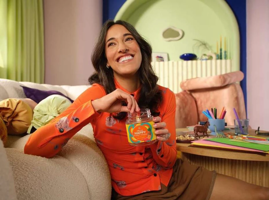 Smiling woman in orange shirt sits on couch, holding a jar of snacks in a casual, indoor setting with colorful decor.