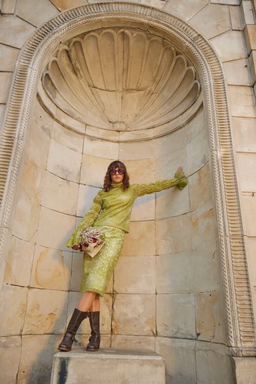 A woman in a green skirt and top poses in front of a stone archway, holding a clutch, wearing brown boots and sunglasses, shot outdoors.
