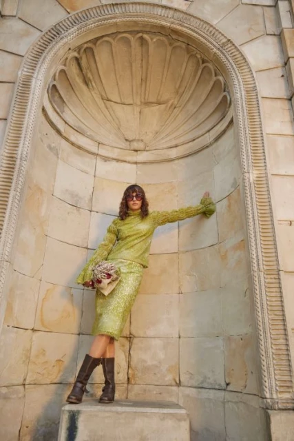 A woman in a green skirt and top poses in front of a stone archway, holding a clutch, wearing brown boots and sunglasses, shot outdoors.