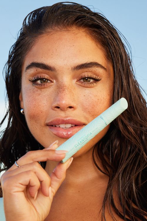 Close-up of a woman with brown hair holding a light blue mascara tube, showcasing beauty and cosmetic products with a serene background.