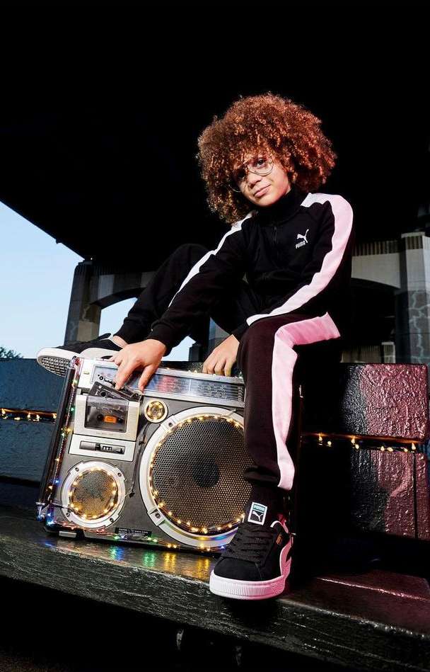 Child model with curly hair poses with boombox, wearing black and white Puma tracksuit, on a rooftop, creating a retro fashion portrait.