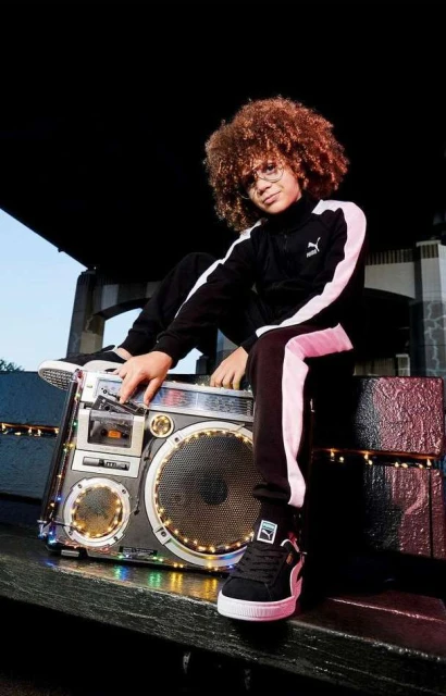Child model with curly hair poses with boombox, wearing black and white Puma tracksuit, on a rooftop, creating a retro fashion portrait.