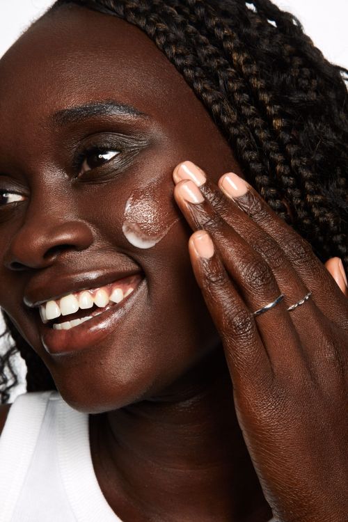 Close-up of a Black woman applying skincare cream to her cheek, photographed by Tory Rust against a plain background.