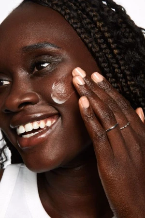 Close-up of a Black woman applying skincare cream to her cheek, photographed by Tory Rust against a plain background.