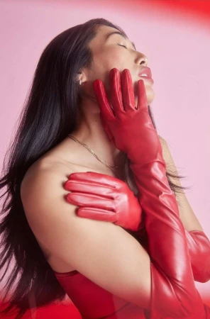 Close-up portrait of a woman wearing bright red opera-length leather gloves with her hands held up to her face against a pink background