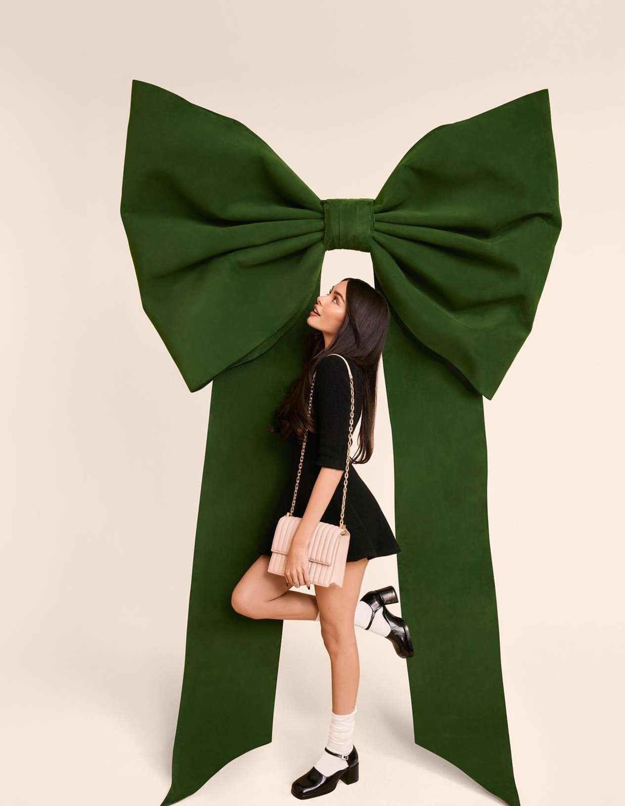 Woman in a black dress and Mary Jane shoes poses with a green ribbon bow against a cream background in a fashion photography studio.