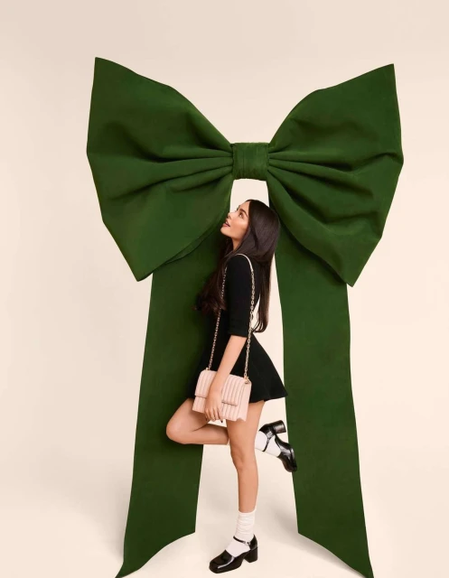 Woman in a black dress and Mary Jane shoes poses with a green ribbon bow against a cream background in a fashion photography studio.