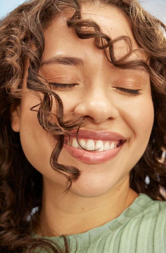 Close-up of a smiling adult woman with curly brown hair and closed eyes. She is wearing a light green top and soft, natural makeup.