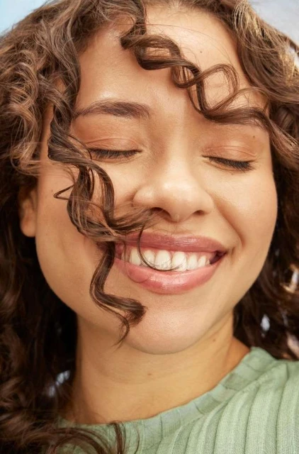 Close-up of a smiling adult woman with curly brown hair and closed eyes. She is wearing a light green top and soft, natural makeup.