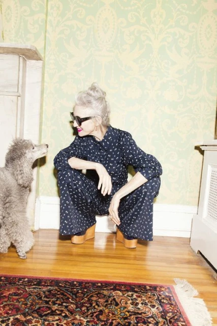 Woman in patterned jumpsuit squats on hardwood floor facing gray poodle in home interior with patterned wallpaper, molding, and rug.