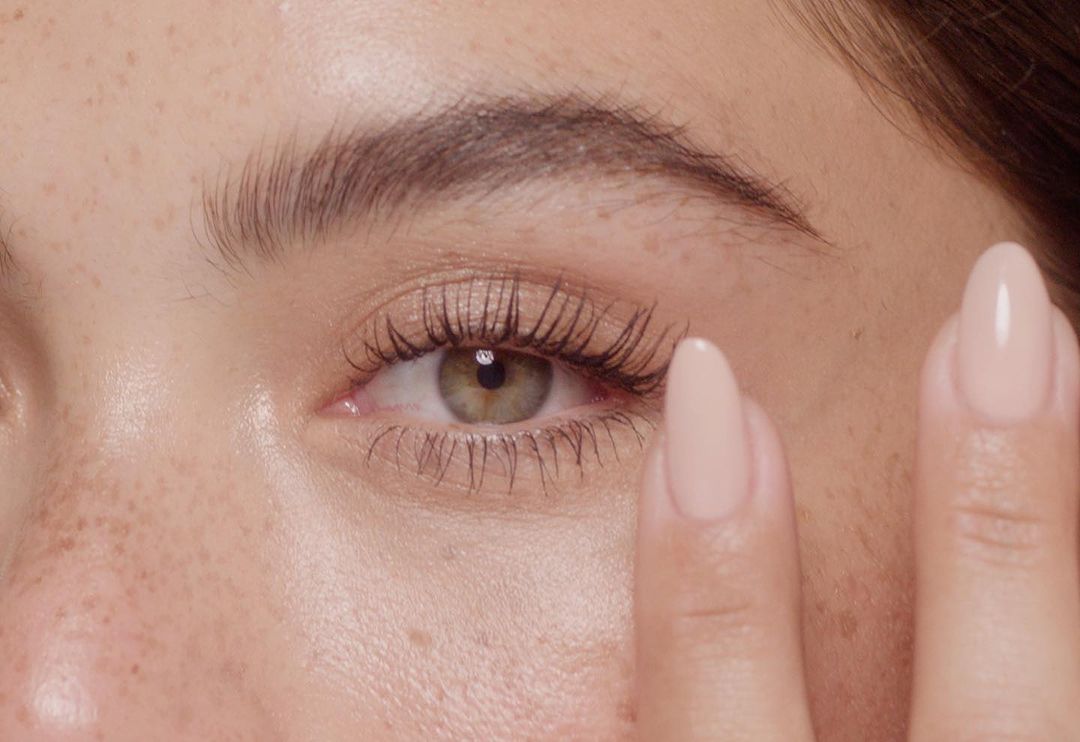 Close-up shot of a woman's eye with long lashes and beige fingernails touching her face, freckles visible, beauty and skincare theme.