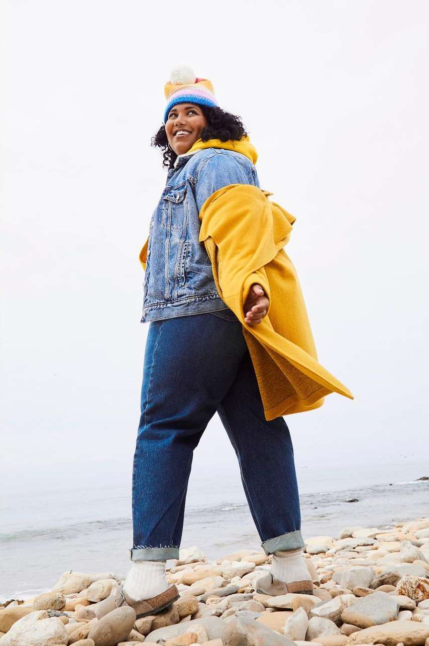 Smiling plus-size woman wearing a beanie and yellow coat standing outdoors on a rocky beach by the ocean.