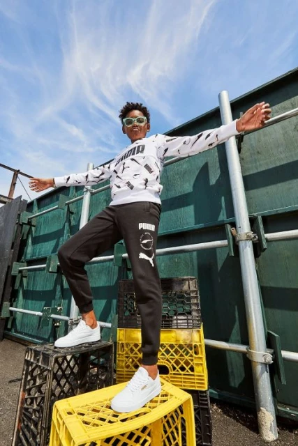 Teenage boy in Puma branded athletic wear strikes a pose on stacked milk crates in outdoor urban setting beneath blue sky.