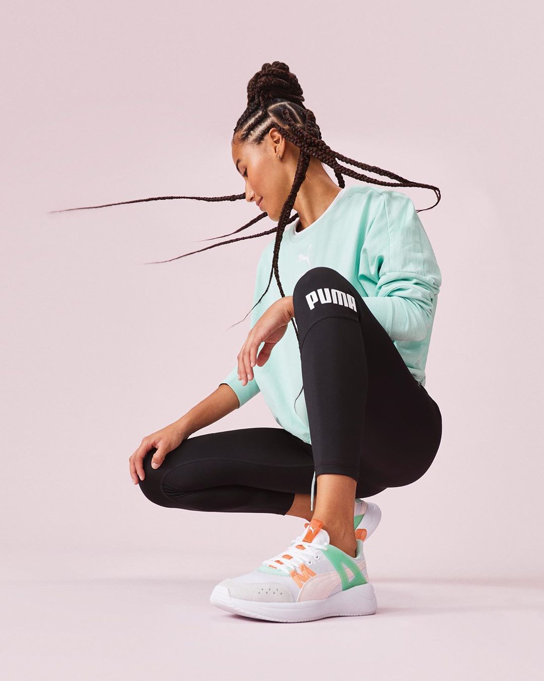 Athletic woman in mint sweatshirt and black leggings poses with braided hair and colorful sneakers against a soft pink backdrop.