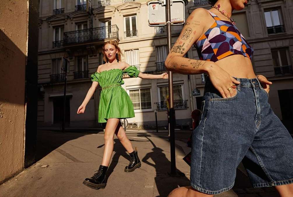 Two women pose in bright sunlight on a European city street. One wears a green dress and the other a patterned top and denim shorts.