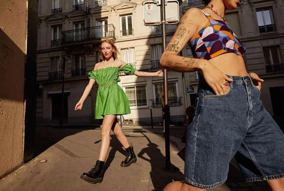 Two women pose in bright sunlight on a European city street. One wears a green dress and the other a patterned top and denim shorts.