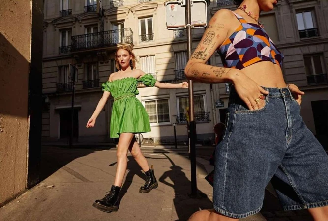 Two women posing on a sunny urban street, one in a green mini dress and another in denim shorts with a patterned crop top.