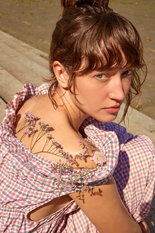 Portrait of a woman with flowers on her shoulder, looking at the camera with a serious expression, wearing a red and white checkered dress.