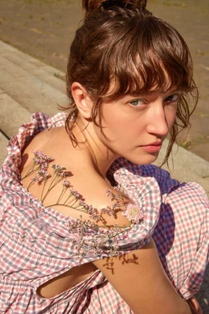 Portrait of a woman with flowers on her shoulder, looking at the camera with a serious expression, wearing a red and white checkered dress.