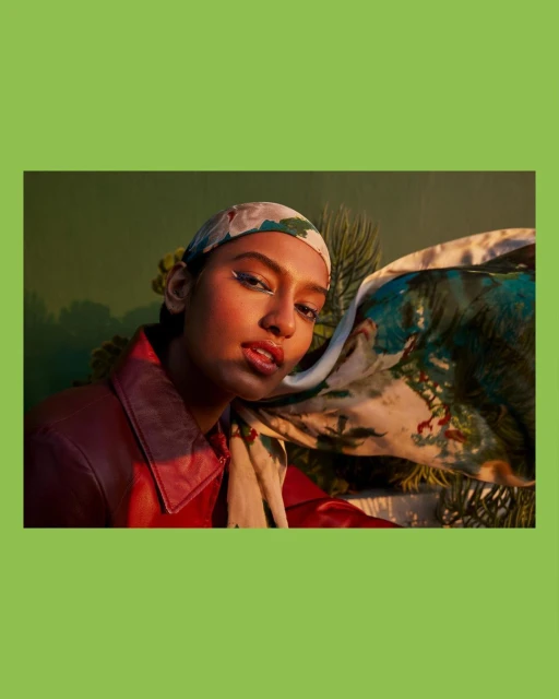 Portrait of a South Asian adult female wearing a headscarf, red jacket, and dramatic eyeliner, posing in a botanically themed setting.