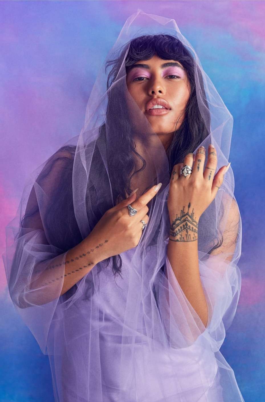 Portrait of a young woman with dark hair and light skin wearing rings, tattoos, and a sheer lilac shawl in front of a vibrant backdrop.