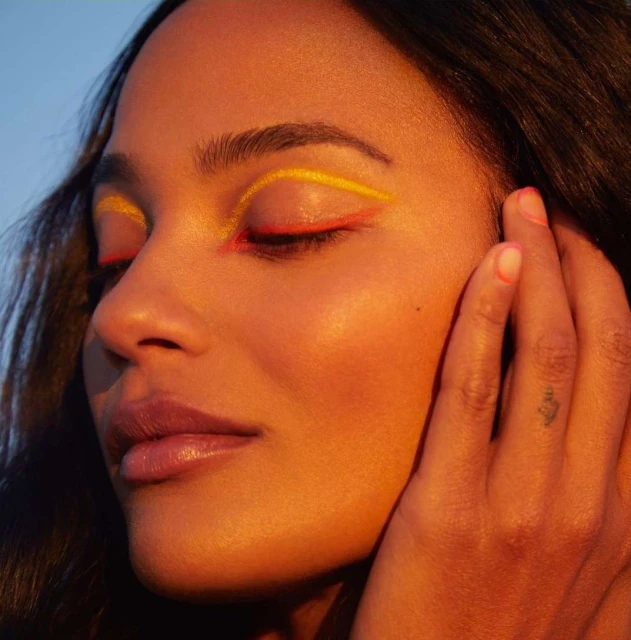 Close-up of a woman with yellow and red eyeliner, eyes closed, and a hand near her face against a bright blue sky background.