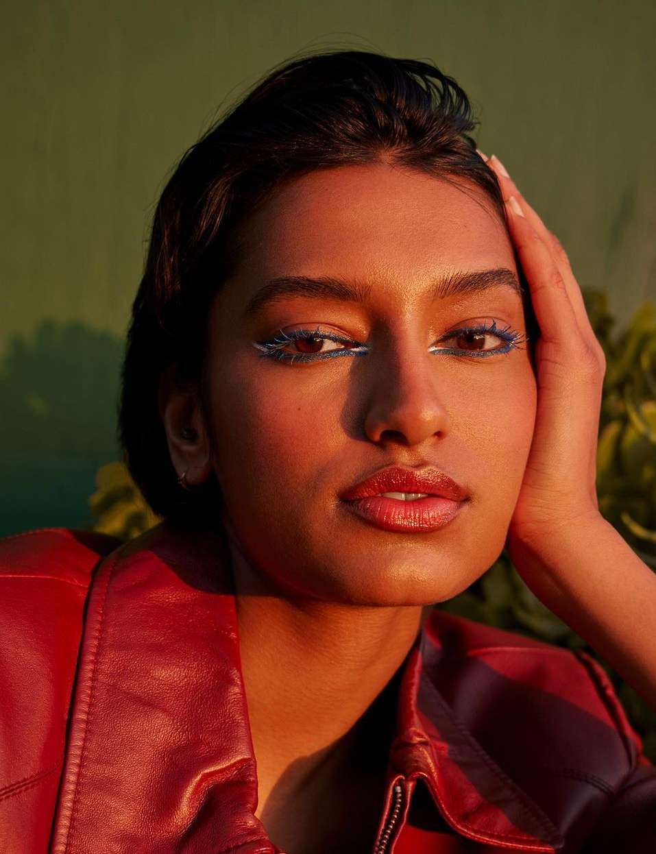 Close-up portrait of an East Asian woman with blue and white eyeliner and a red leather jacket, posing with her hand on her head.