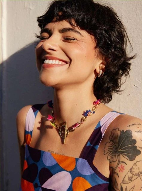 Smiling woman in a colorful print top and floral necklace outside in dappled light. She has short dark hair and visible tattoos.