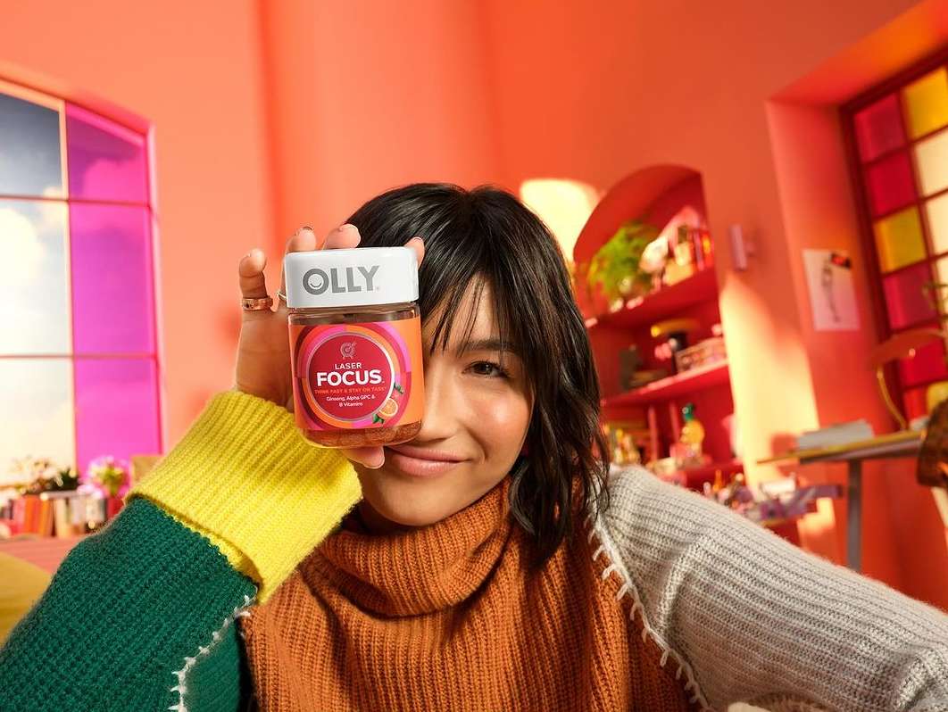 Woman holding Olly Laser Focus supplement in orange room with colorful bookshelf. Close up of female smiling, wearing a turtleneck sweater.