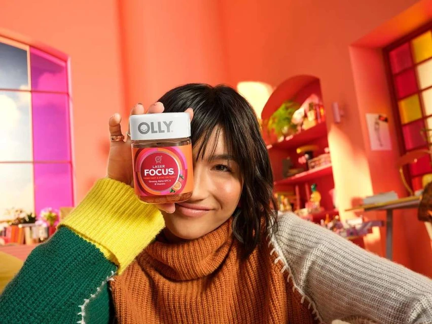Woman holding Olly Laser Focus supplement in orange room with colorful bookshelf. Close up of female smiling, wearing a turtleneck sweater.