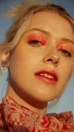 Close-up portrait of a blonde woman with glossy pink eyeshadow and red lips wearing a patterned red scarf under golden sunlight.