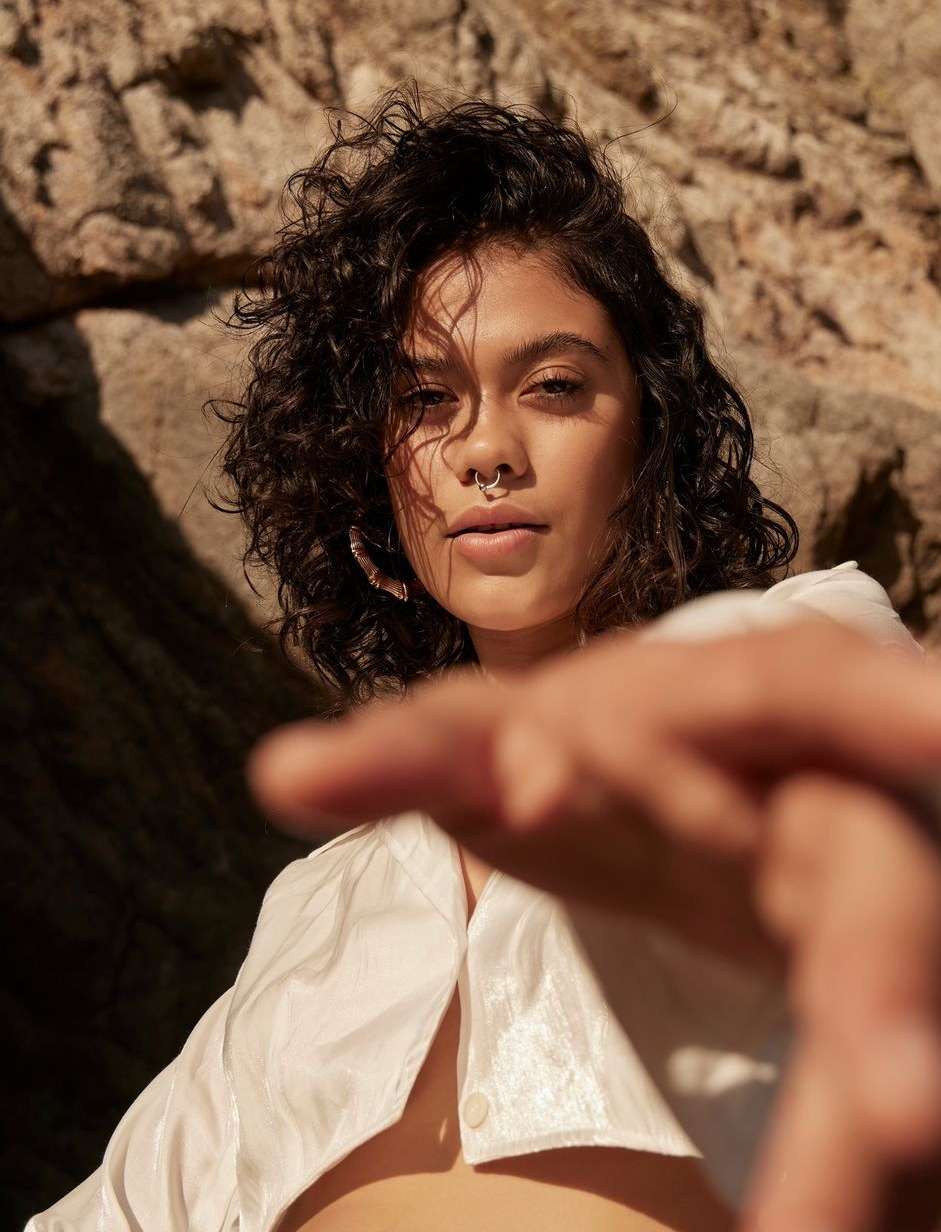 Close-up shot of a young woman with curly dark hair, a nose ring, and a white top, posing in front of a rock formation with dramatic lighting.