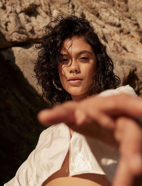 Close-up shot of a young woman with curly dark hair, a nose ring, and a white top, posing in front of a rock formation with dramatic lighting.
