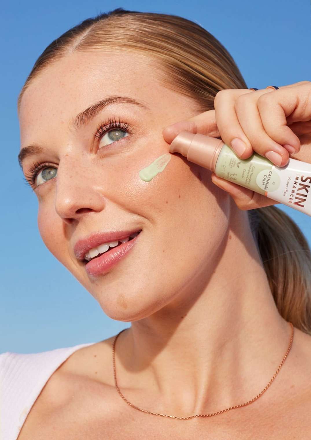 Smiling woman applying skincare product on her face outdoors against a blue sky backdrop, showcasing beauty and cosmetic usage.