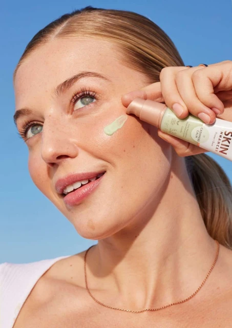 Smiling woman applying skincare product on her face outdoors against a blue sky backdrop, showcasing beauty and cosmetic usage.