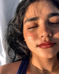 Close-up portrait of a South Asian woman with closed eyes, glossy dark red lips, and two small dots on her cheek under warm sunlight.