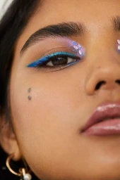 Extreme close-up of a person with vibrant blue eyeliner, iridescent purple eyeshadow, and face gems captured by Tory Rust