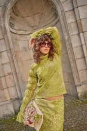 Model in green patterned top and sunglasses holding textured bag standing before stone architecture