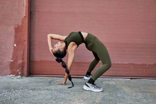 Woman in olive green athletic set performing a backbend on pavement against a reddish brown wall