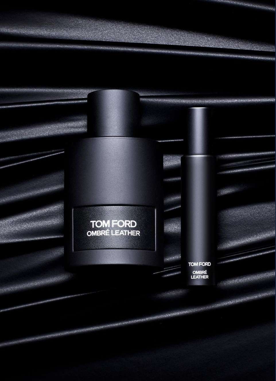Tom Ford Ombre Leather fragrance still life. A bottle and travel spray are staged on a black satin textile. Perfume and luxury aesthetic.