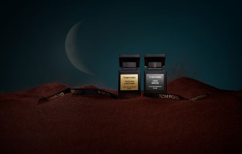 Two Tom Ford fragrance bottles, Tuscan Leather and Oud Wood, sit on rust-colored sand with a black Tom Ford ribbon and crescent moon backdrop.