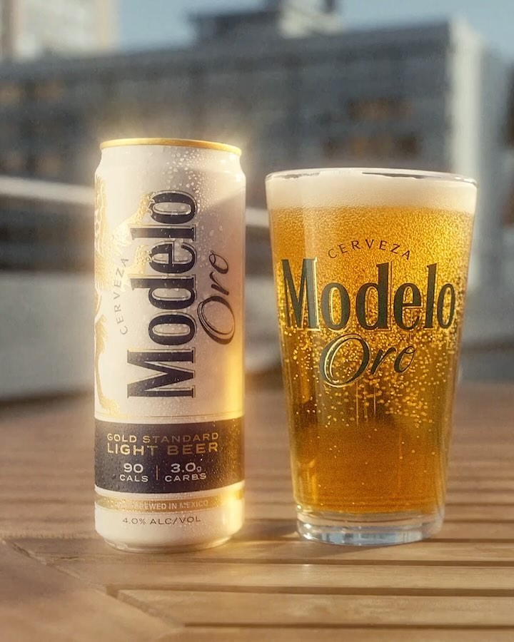 Modelo Oro beer can and glass, featuring gold accents and condensation, set on a wooden surface, with a blurred outdoor background.