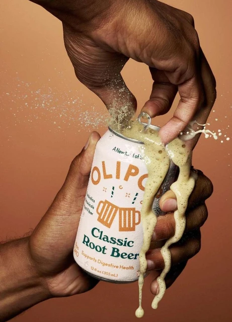 Close-up studio shot of a hand opening a can of Olipop Classic Root Beer, causing it to overflow, set against a warm brown background.